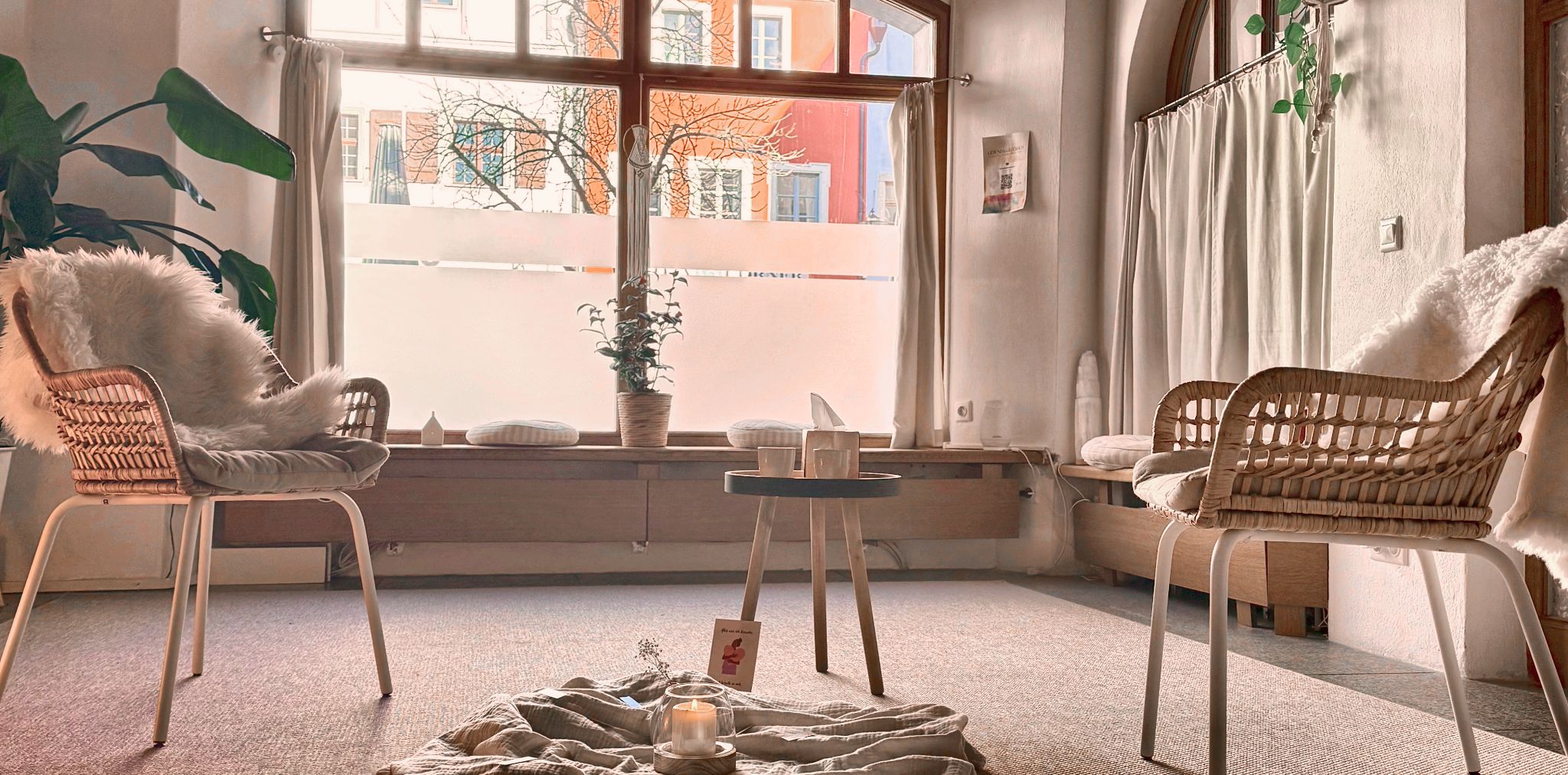 Der Praxisraum im Gegenlicht: zwei Korbstühle am großen Bogenfenster, davor eine Kerze auf einer Decke am Boden